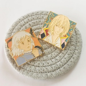 May include: Two enamel pins featuring anime characters. One pin depicts a blonde-haired character in a gold frame with a detailed background. The other pin shows a character with white hair in a blue dress, set against an orange backdrop.
