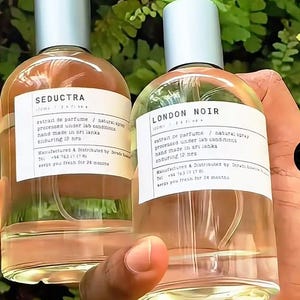 May include: Two clear glass perfume bottles with silver caps. One bottle is labeled "SEDUCTRA", the other "LONDON NOIR". Both bottles have white labels with black text. The bottles are held in front of a green leafy background.