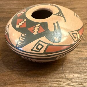 May include: A hand-painted, oval-shaped pottery vessel with a central opening. The beige-colored pot features a black, red, and green geometric design, including stylized bird and zigzag patterns. The rim and base are also decorated with black lines.