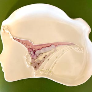 May include: A white ceramic decorative dish with a profile of a woman's face. The dish features a design of pink, purple, and gold accents. The dish has a wavy edge and is set against a green background.