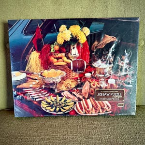 May include: A Springbok jigsaw puzzle box featuring a tailgate party scene with a football, silver serving dishes, hot dogs, sliced ham, fruit, and yellow flowers. The box includes the text: AN AUTHENTIC INTERLOCKING JIGSAW PUZZLE by Springbok OVER 500 PIECES.