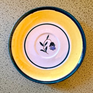 May include: A decorative ceramic saucer with a yellow rim and a dark blue edge. The center is white with a green and black floral design and a purple flower. The saucer is round.