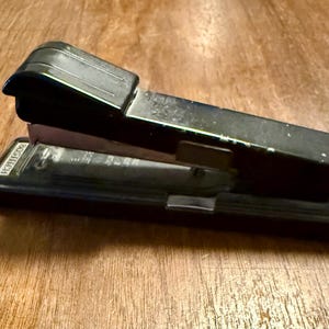 May include: A black Bostitch stapler on a wooden surface. The stapler is made of metal and has a lever-style design. The word "Bostitch" is visible on the side of the stapler.