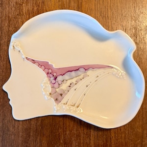 May include: A white ceramic decorative tray in the shape of a woman's profile. The tray features a design of pink, purple, and gold accents, resembling a waterfall or flowing hair. The tray has a scalloped edge and is set on a wooden surface.