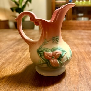 May include: A small, vintage pink ceramic pitcher with a curved handle and a spout. The pitcher features a floral design with green leaves and a red flower. The base is a light cream color.