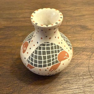 May include: A small, off-white ceramic vase with a wide opening and a rounded body. The vase is decorated with a repeating pattern of orange turtles and black grid designs. Small orange and black dots are scattered across the surface.