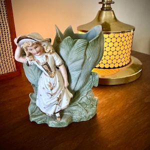 May include: A vintage porcelain figurine of a girl in a white dress and hat, leaning against a green tulip-shaped vase. The vase has a light blue interior and gold accents. A brass lamp is in the background.