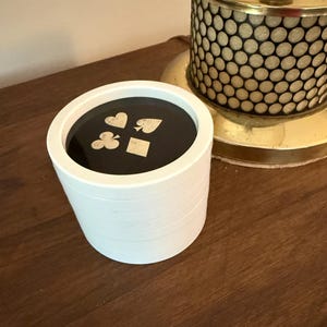 May include: A white, cylindrical playing card holder with a black interior. The top features gold card suit symbols: hearts, spades, clubs, and diamonds. The holder is on a wooden surface, with a gold lamp in the background.
