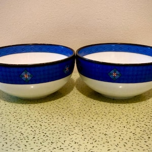 May include: Two white and blue ceramic bowls. The bowls have a white interior and a blue exterior with a decorative pattern. The rim of the bowls is also blue with a decorative pattern. The bowls are sitting on a speckled surface.