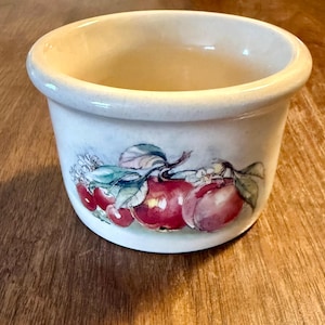 May include: A small, off-white ceramic bowl with a rounded rim. The bowl features a painted design of red apples with green leaves and white blossoms. The bowl is sitting on a wooden surface.