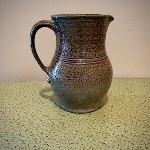 May include: A stoneware pitcher with a textured surface and a dark brown and gray color scheme. The pitcher has a curved handle and a spout for pouring. The design includes two horizontal bands around the body. This rustic pitcher is suitable for serving beverages.