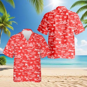 Puede incluir: Camisa roja de manga corta con un estampado tropical blanco, que incluye palmeras, barcos y la palabra "Nebraska". La camisa también presenta la letra "N".