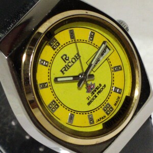 May include: A vintage Ricoh automatic wristwatch with a bright yellow face. The watch features a silver-tone case, a black leather band, and the text "21 Jewels Shock Proof Japan Made". The watch face displays Roman numerals and the Ricoh logo.