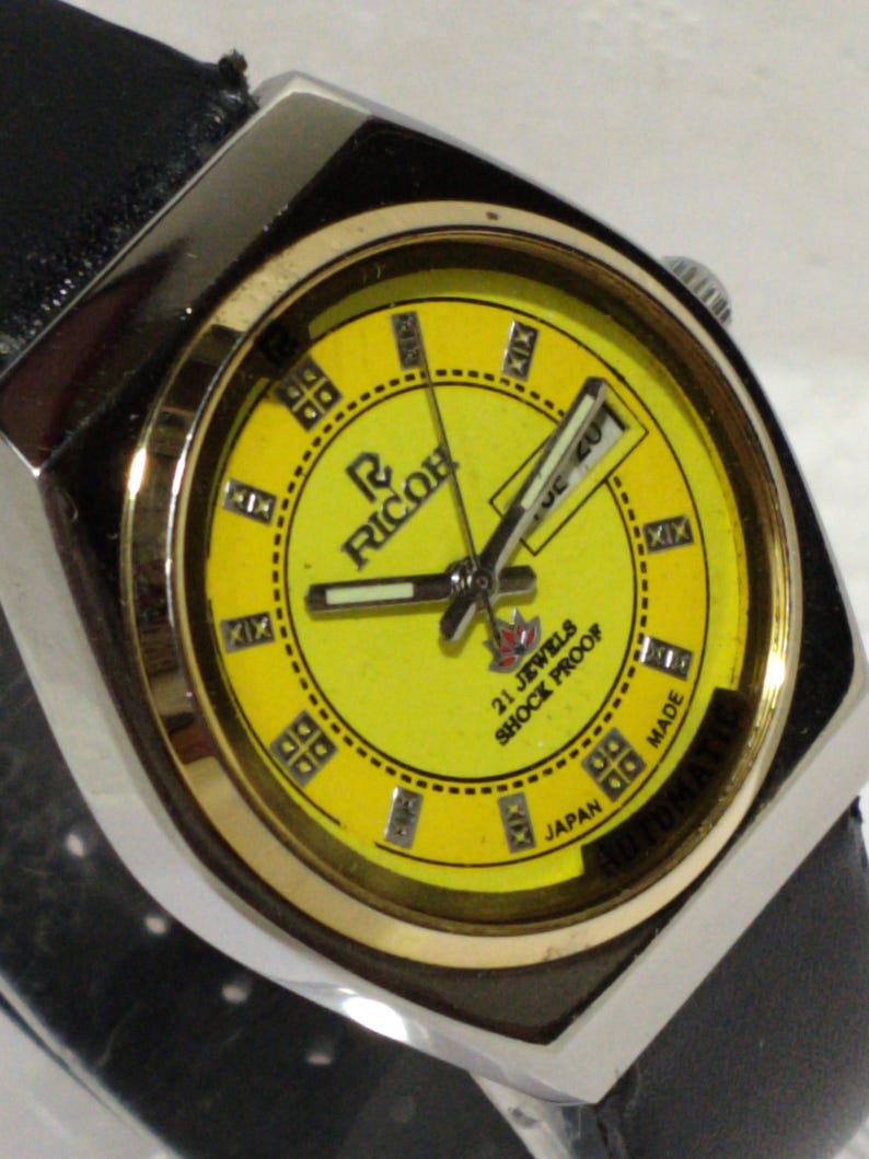 May include: A vintage Ricoh automatic wristwatch with a bright yellow face. The watch features a silver-tone case, a black leather band, and the text "21 Jewels Shock Proof Japan Made". The watch face displays Roman numerals and the Ricoh logo.