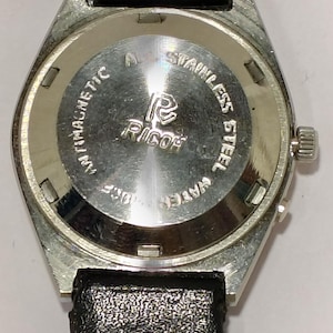 May include: A vintage Ricoh wristwatch with a stainless steel back and a black leather strap. The watch face is silver with the Ricoh logo and the words "Antimagnetic" and "Stainless Steel". The watch is water resistant.
