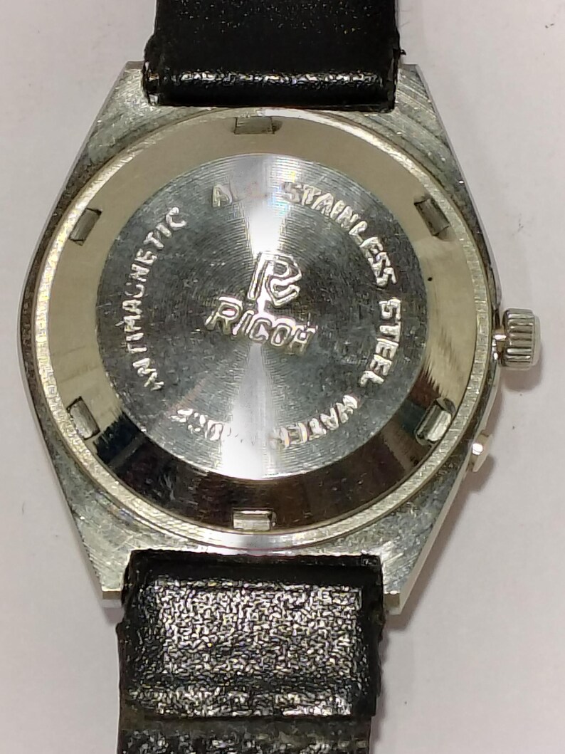 May include: A vintage Ricoh wristwatch with a stainless steel back and a black leather strap. The watch face is silver with the Ricoh logo and the words "Antimagnetic" and "Stainless Steel". The watch is water resistant.