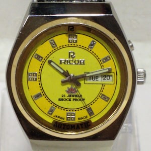 May include: Vintage Ricoh automatic watch with a yellow face, gold-tone bezel, and a black leather strap. The watch face displays the day and date, with the text "21 Jewels Shock Proof" and "Japan Made".