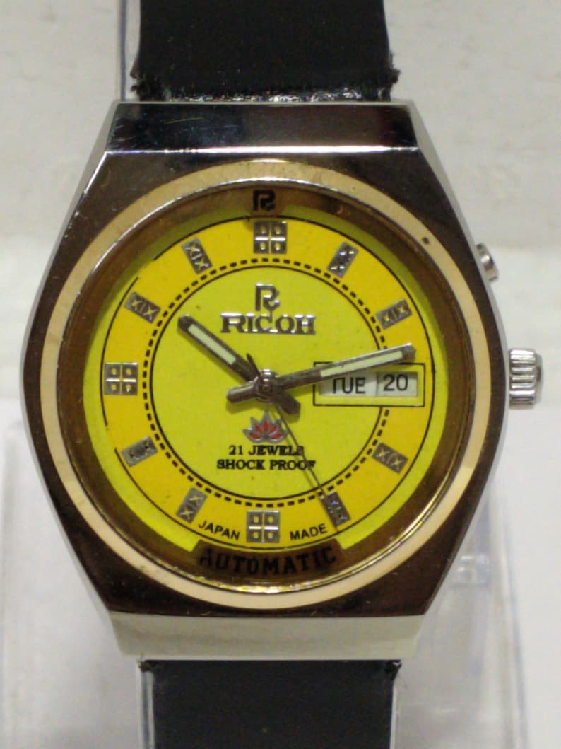 May include: Vintage Ricoh automatic watch with a yellow face, gold-tone bezel, and a black leather strap. The watch face displays the day and date, with the text "21 Jewels Shock Proof" and "Japan Made".