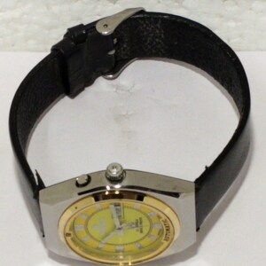 May include: A wristwatch with a silver-tone case and a gold-colored bezel. The watch face is yellow with black numerals and hands. It has a black leather band. The watch is an automatic model.