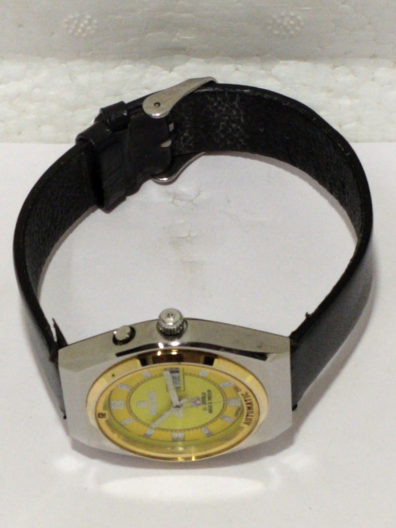May include: A wristwatch with a silver-tone case and a gold-colored bezel. The watch face is yellow with black numerals and hands. It has a black leather band. The watch is an automatic model.