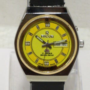 May include: A vintage Ricoh automatic watch with a yellow face and a black leather strap. The watch has a silver-colored case and features the day and date. The watch face displays the text "RICOH", "21 JEWELS", "SHOCK PROOF", "JAPAN MADE", and "AUTOMATIC".