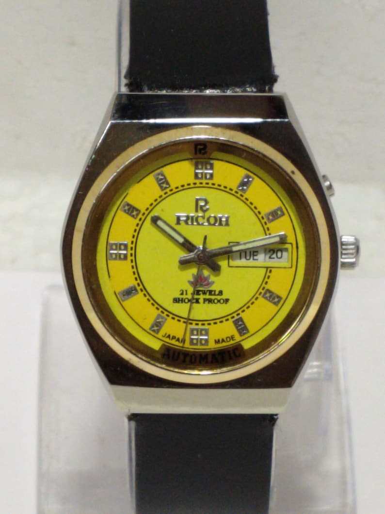May include: A vintage Ricoh automatic watch with a yellow face and a black leather strap. The watch has a silver-colored case and features the day and date. The watch face displays the text "RICOH", "21 JEWELS", "SHOCK PROOF", "JAPAN MADE", and "AUTOMATIC".