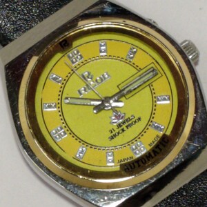 May include: A vintage wristwatch with a yellow face and silver-tone case. The watch face features the brand name "Ricoh", the text "21 Jewels Shock Proof", and the word "Automatic". The watch has a black leather band.