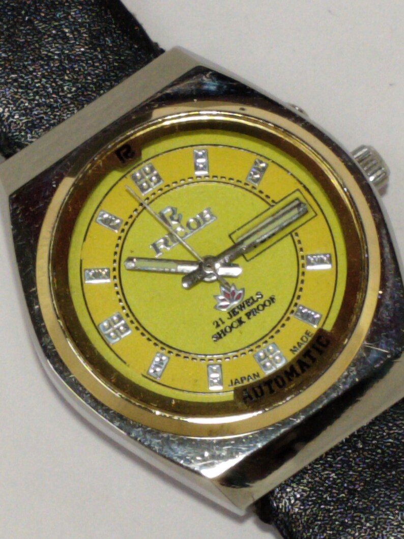 May include: A vintage wristwatch with a yellow face and silver-tone case. The watch face features the brand name "Ricoh", the text "21 Jewels Shock Proof", and the word "Automatic". The watch has a black leather band.