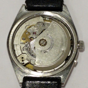 May include: A vintage wristwatch with a silver-colored case and a black leather band. The watch's back is open, revealing the intricate mechanical movement with gears, springs, and a rotor. The watch is a classic timepiece.