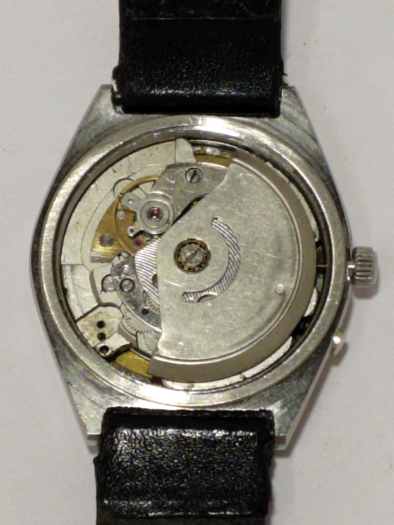 May include: A vintage wristwatch with a silver-colored case and a black leather band. The watch's back is open, revealing the intricate mechanical movement with gears, springs, and a rotor. The watch is a classic timepiece.