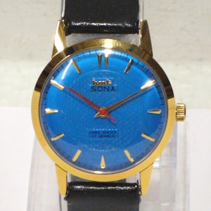 Könnte beinhalten: Eine Armbanduhr mit einem goldfarbenen runden Gehäuse und einem leuchtend blauen Zifferblatt. Das Zifferblatt zeigt goldfarbene Stundenmarkierungen, einen roten Sekundenzeiger und den Text HMT SONA PARA SHOCK 17 JEWELS.