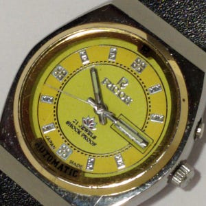 May include: A vintage wristwatch with a yellow face and silver-tone case. The watch face features the text "RICOH", "21 JEWELS", "SHOCK PROOF", and "AUTOMATIC". The watch has a black leather band.