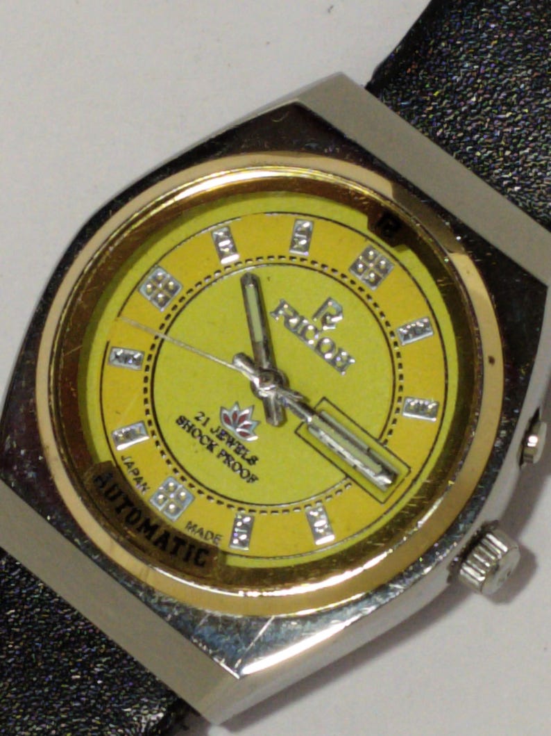 May include: A vintage wristwatch with a yellow face and silver-tone case. The watch face features the text "RICOH", "21 JEWELS", "SHOCK PROOF", and "AUTOMATIC". The watch has a black leather band.