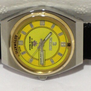 May include: A vintage wristwatch with a yellow face and gold-tone bezel. The watch has a silver-tone case and a black leather strap. The face features the brand name "Ricoh" and the words "Automatic", "Japan", and "21 Jewels Shock Proof".
