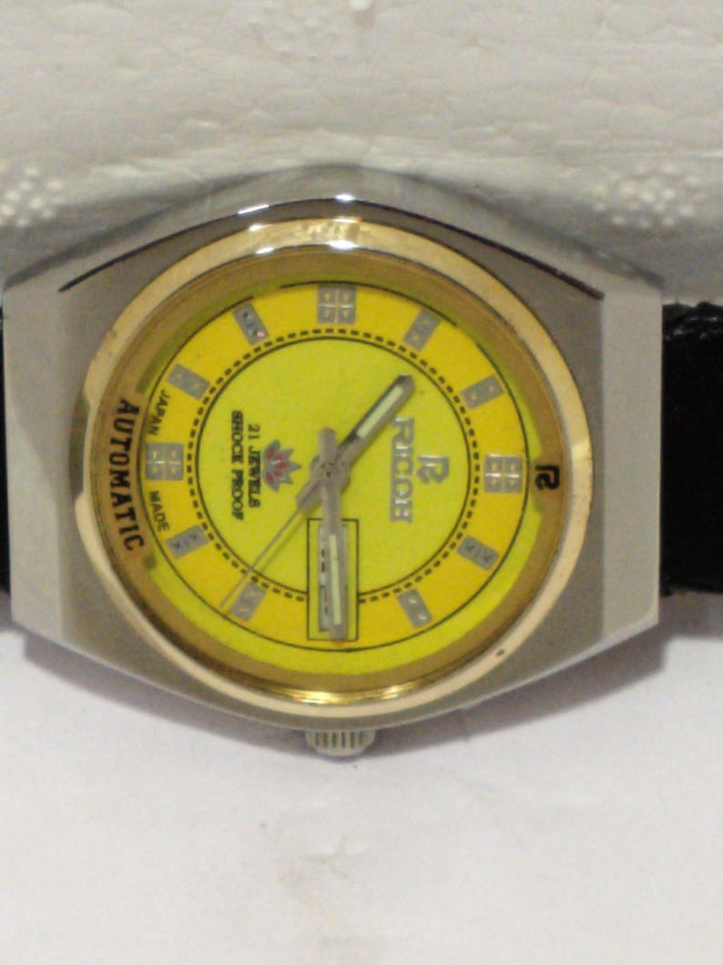 May include: A vintage wristwatch with a yellow face and gold-tone bezel. The watch has a silver-tone case and a black leather strap. The face features the brand name "Ricoh" and the words "Automatic", "Japan", and "21 Jewels Shock Proof".