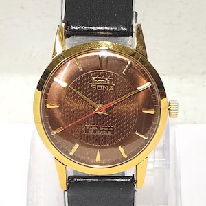 May include: A gold-tone wristwatch with a brown textured dial, gold hour markers, and a black leather strap. The dial features the text HMT SONA, PARA SHOCK, and 17 JEWELS. It has gold hour and minute hands with a thin red second hand.