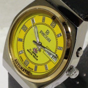May include: A vintage Ricoh automatic wristwatch with a yellow face and a black leather band. The watch has a silver-tone case with gold-tone accents and a date display. The watch face features the text "21 Jewels Shock Proof" and "Automatic Japan Made".
