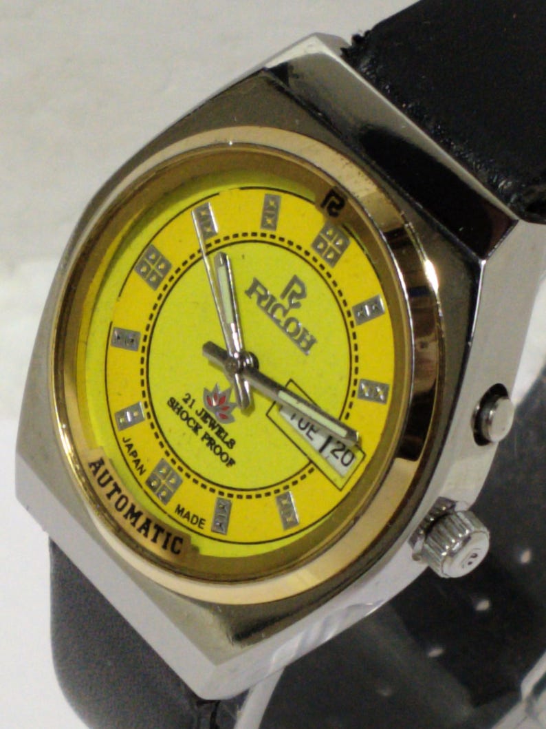 May include: A vintage Ricoh automatic wristwatch with a yellow face and a black leather band. The watch has a silver-tone case with gold-tone accents and a date display. The watch face features the text "21 Jewels Shock Proof" and "Automatic Japan Made".