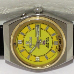 May include: A vintage Ricoh automatic wristwatch with a yellow dial and a silver-tone case. The watch face displays the day of the week and features the text "21 Jewels Shock Proof Japan Made". The watch has a black leather strap.