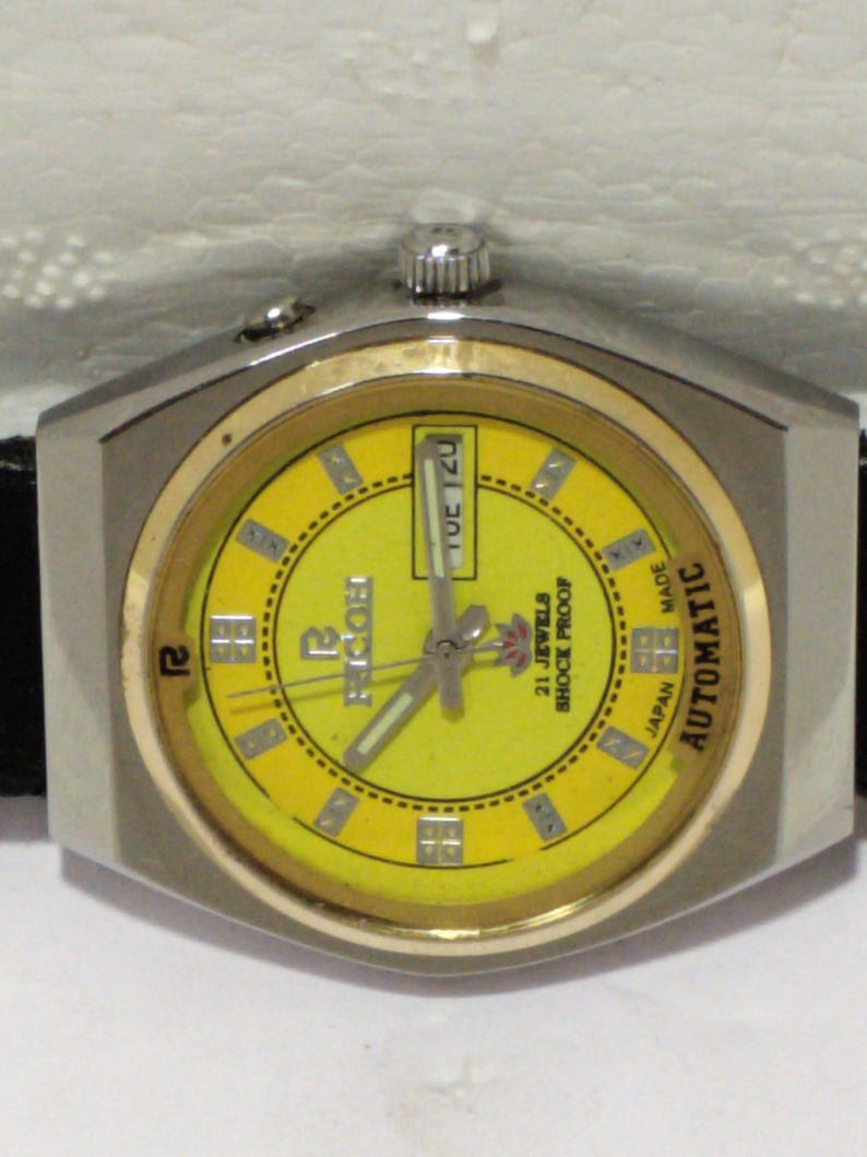 May include: A vintage Ricoh automatic wristwatch with a yellow dial and a silver-tone case. The watch face displays the day of the week and features the text "21 Jewels Shock Proof Japan Made". The watch has a black leather strap.