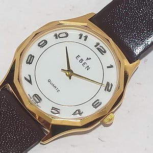 Puede incluir: Un reloj de cuarzo dorado con esfera blanca, números negros y la marca "EBEN". El reloj tiene una correa de cuero marrón. La esfera del reloj tiene aproximadamente 3,2 cm de diámetro.