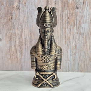 May include: A bronze-colored bust of an Egyptian pharaoh. The statue features a detailed headdress, ornate collar, and crossed scepters at the base. The figure has a serene expression and is a decorative item.