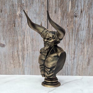 May include: A bronze-colored bust of a demonic figure with large horns, a snarling expression, and muscular torso. The sculpture is mounted on a pedestal base and set against a wooden background.