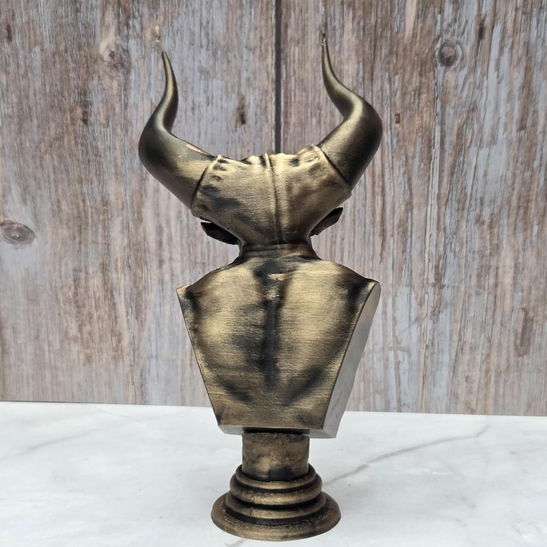 May include: A bronze-colored bust of a horned figure, viewed from the back. The sculpture features a detailed depiction of the figure's back, shoulders, and the base of the horns. The bust is mounted on a cylindrical pedestal.