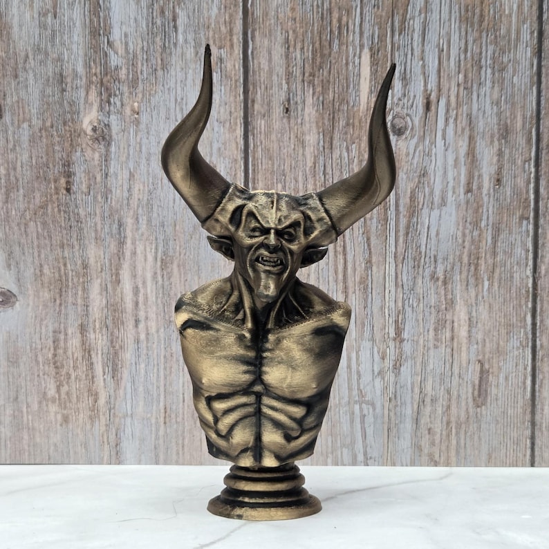 May include: A bronze-colored bust of a demonic figure with large horns, a snarling expression, and a muscular torso. The sculpture is mounted on a pedestal base, showcasing intricate details and textures. The bust is set against a wooden background.