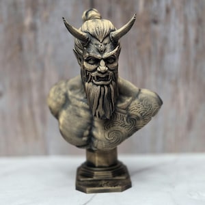May include: A bronze-colored bust of a horned demon with a long beard and intricate facial details. The sculpture features a detailed tattoo on the shoulder and is mounted on a hexagonal base. The demon has a stern expression.