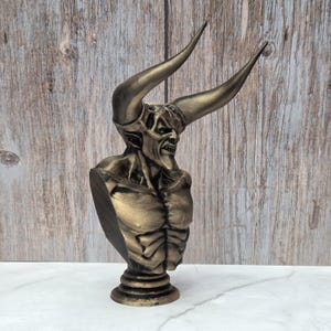May include: A bronze-colored bust of a horned demon with a menacing expression. The sculpture features large, curved horns and detailed musculature. The bust is mounted on a round, tiered base, showcasing a gothic or fantasy theme.