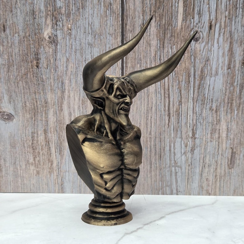 May include: A bronze-colored bust of a horned demon with a menacing expression. The sculpture features large, curved horns and detailed musculature. The bust is mounted on a round, tiered base, showcasing a gothic or fantasy theme.