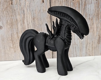 May include: A black figurine of a hybrid creature, combining the features of a pony and an alien. The figure has a pony-like body with a long, curved tail and a head resembling the Xenomorph from the Alien film series. The figurine is displayed on a white surface.