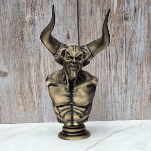 May include: A bronze-colored bust of a horned demon with a menacing expression. The sculpture features detailed musculature and a textured finish, mounted on a circular base. The horns curve upwards, adding to the imposing presence of the figure.